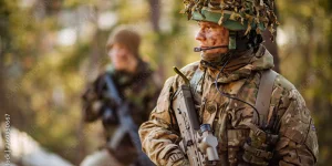 Soldier in camouflage gear with a headset and rifle, standing outdoors with another soldier blurred in the background.