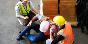 Two construction workers wearing safety vests and helmets appear to be assisting a person lying on the floor at a construction site, with a stack of bricks nearby.