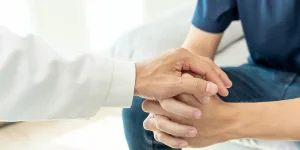 Person in a white coat gently holding the hands of a person in a blue shirt during a comforting visit or medical consultation.