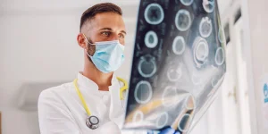 A male doctor wearing a face mask and stethoscope examining brain scan images on a lightbox in a medical setting.