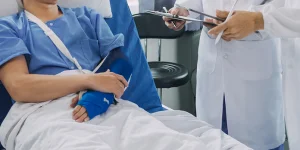 A patient in a hospital bed with a blue hospital gown, arm in a cast, being attended to by a healthcare professional with a stethoscope and clipboard.