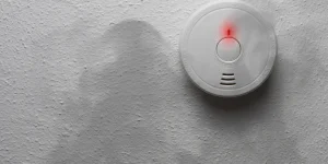 A smoke detector with a red warning light is mounted on a textured white ceiling.