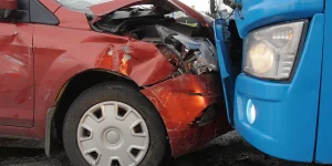 Red car and blue truck collided at front, with significant damage to the red car's front end, crumpled hood, and partially detached bumper. Debris scattered on the road.