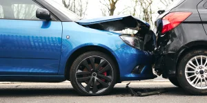 Blue car with front-end damage connecting with the rear of a black SUV in a car accident on a road.