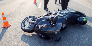 A black motorcycle lies on its side on the road with orange traffic cones nearby, and people standing around it, possibly after an accident.