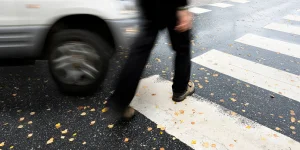 Person crossing a street at a crosswalk with falling leaves, near a moving vehicle on a rainy day.
