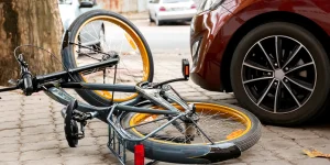 A black and yellow bicycle is lying on its side on a sidewalk next to a parked red car. The bike's wheels and frame are visible, with some leaves nearby.