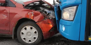 Red car front-end collision with a blue bus, showing significant damage to the car's front quarter panel and bumper. Debris on the road suggests a recent crash.