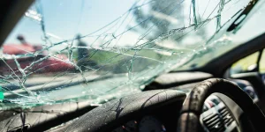 Broken windshield with multiple cracks and shattered glass visible inside a car. The steering wheel and dashboard are partially shown.