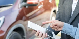 Person in a suit using a tablet beside a brown car, with another person pointing at the tablet.