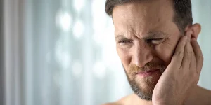Man with a beard holding his head in pain or discomfort, expression showing distress or concern, with a blurred light background.