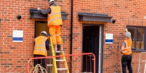 Three construction workers in orange safety vests and helmets install or repair a black box on a brick building exterior. One worker is on a ladder, another on the ground, and a third standing nearby.