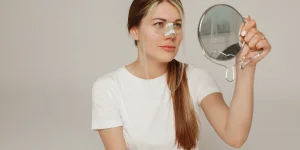 Woman with a nasal tube, looking into a handheld mirror, wearing a white t-shirt, with a neutral background.