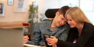 A teenage boy with a disability sits in a wheelchair, showing a smartphone to a smiling adult woman in an indoor setting with a bright background.