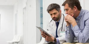 A doctor and a man are sitting in a hospital corridor, looking at a tablet together with focused expressions, possibly discussing medical information.