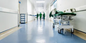 Empty hospital corridor with a hospital bed on wheels and healthcare staff walking in the distance. Bright lighting and clean, sterile environment.