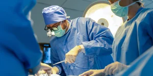 Surgeons in blue scrubs and masks perform an operation in a brightly lit operating room.