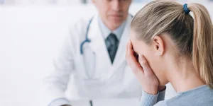 A woman with a ponytail covering her face, sitting in a medical setting, with a doctor in the background holding a clipboard and wearing a stethoscope.