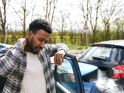 Man examining his neck next to a blue car with smoke coming from the hood in a parking lot, with trees and open sky in the background.