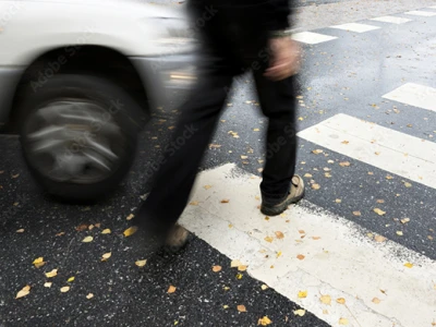 A person crossing a street at a crosswalk as a white car passes by, with fallen yellow leaves scattered on the wet asphalt.