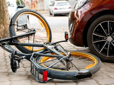 A black bicycle with yellow accents lies on its side on a brick sidewalk near a parked red car, with trees and other cars visible in the background.