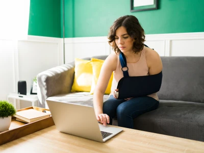 A woman with a curly bob hairstyle sits on a gray sofa in a brightly lit living room, using a laptop with her right arm in a blue sling.