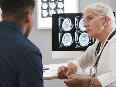A female healthcare professional, possibly a doctor, discusses brain scan images with a male patient in a medical office. The monitor displays multiple brain MRI scans.