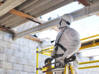 A construction worker in white protective gear and a helmet is working on a building site, standing on a yellow scaffold with concrete blocks and wooden beams overhead.