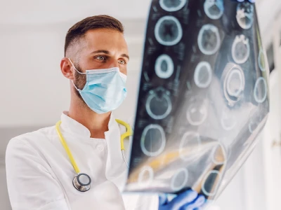 A male doctor wearing a face mask and stethoscope examining brain scan images on a lightbox in a medical setting.