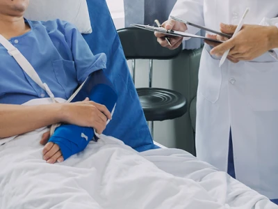 A patient in a hospital bed with a blue hospital gown, arm in a cast, being attended to by a healthcare professional with a stethoscope and clipboard.