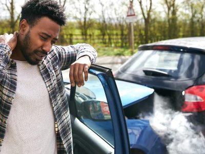 A man with curly black hair and a beard appears distressed while standing outside next to a blue car involved in a rear-end collision with a black vehicle, as smoke or steam rises from the point of impact.