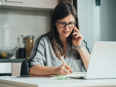 A woman with dark hair, glasses, and a grey shirt is sitting at a desk, talking on the phone, writing in a notebook, with a laptop and some stationery on the desk.