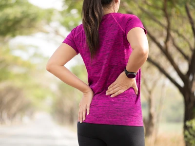 A woman in a pink athletic shirt with black leggings, standing outdoors on a tree-lined pathway, holding her lower back with one hand, and wearing a smartwatch.