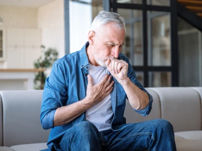 An elderly man with gray hair and a beard sits on a beige sofa, clutching his chest and coughing into his fist, appearing to experience chest pain or discomfort.