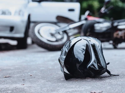 A black and white motorcycle helmet lying on the ground in front of a car and a motorcycle, which are slightly blurred in the background.