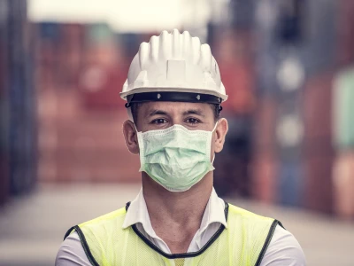 A man wearing a white safety helmet, a face mask, a white shirt, and a yellow safety vest stands outdoors with a blurred industrial background.