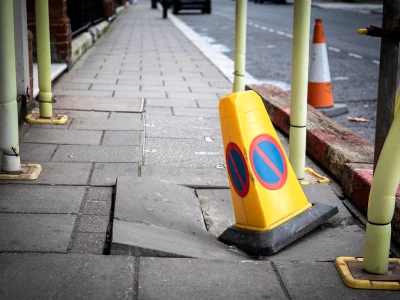 A yellow traffic cone with blue and red no-parking symbols leaning on a sidewalk with broken pavement, surrounded by safety barriers on a city street.