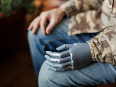 A person in camouflage clothing with a prosthetic robotic hand resting on their lap, with another hand on their knee. The background is blurred.