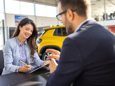A woman smiling and taking notes at a desk while talking to a man in a suit, in a car dealership or showroom with a yellow vehicle in the background.