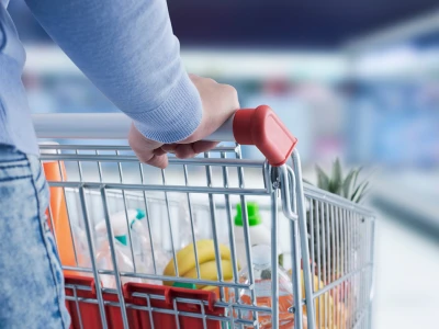 A person wearing a light blue sweater pushes a shopping cart filled with groceries in a store aisle.