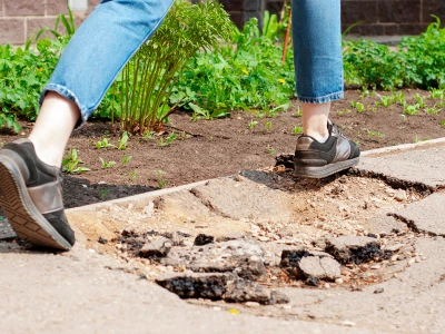 Person repairing a broken sidewalk with visible cracks and loose stones, wearing black sneakers and blue jeans, next to a garden with green plants.