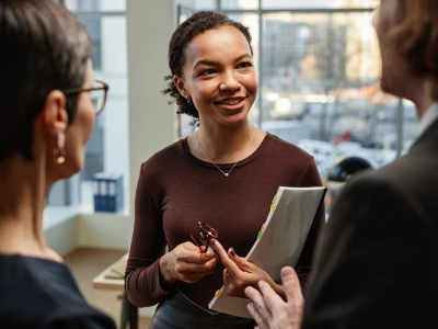 A woman smiling and holding a folder with sticky notes, engaged in conversation with two people in an office setting with large windows and city views.