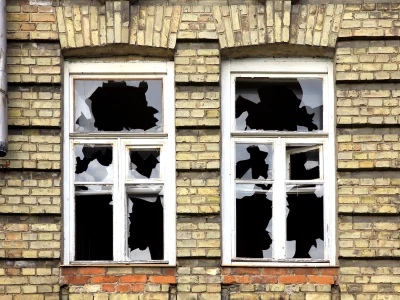 Two broken windows with shattered glass in a brick wall building, with holes and jagged edges in the glass panes, and a metal drainpipe on the left side.