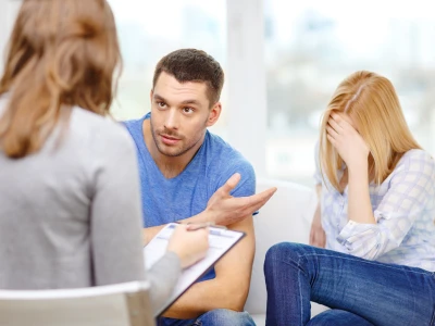 A young man in a blue shirt appears to be speaking to a woman, with a woman on the right covering her face with her hand, possibly upset or distressed.