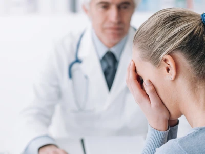 A woman with a ponytail covering her face, sitting in a medical setting, with a doctor in the background holding a clipboard and wearing a stethoscope.