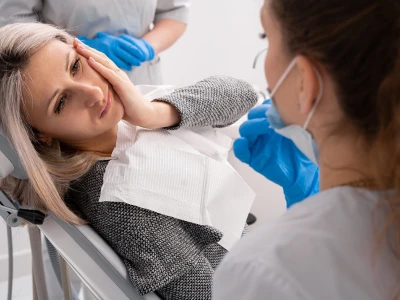 A woman with blonde hair lying in a dental chair with her hand on her temple, while a female healthcare worker wearing masks and blue gloves examines her mouth.