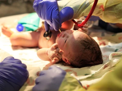 Newborn baby lying on a blanket with medical personnel tending to it, wearing purple gloves, and using medical instruments during a delivery.