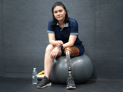 A young woman with a prosthetic leg and a tiger-patterned knee sits on a large exercise ball, with a water bottle and gym equipment nearby, against a dark wall.