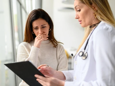 A woman with a worried expression covers her mouth with her hand while talking to a healthcare professional who holds a clipboard, in a clinical setting.