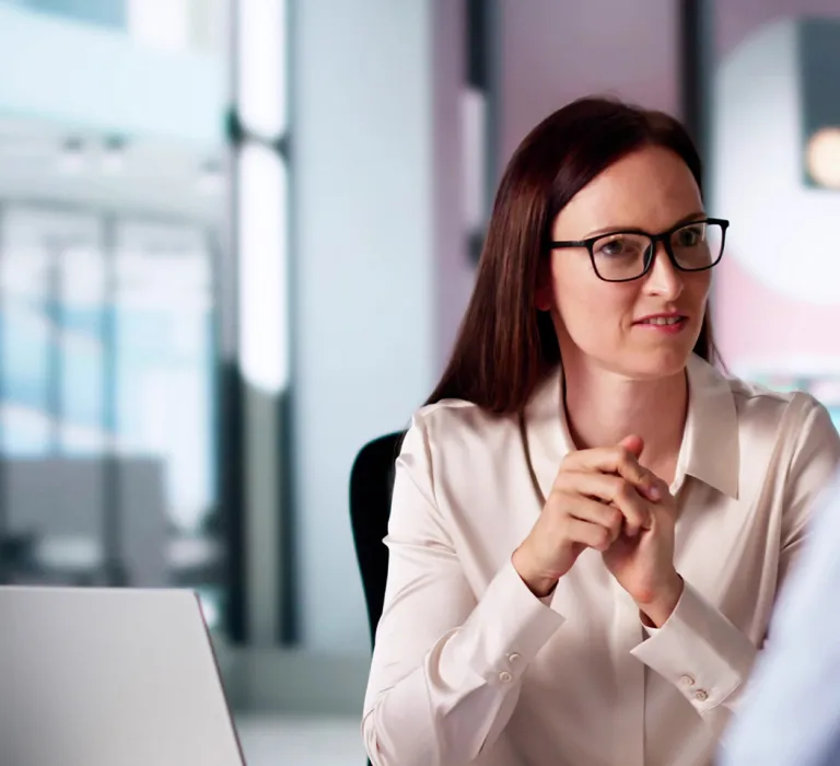 A woman with brown hair, glasses, and a white blouse sits at a desk with hands clasped, engaged in a conversation with a person facing away.